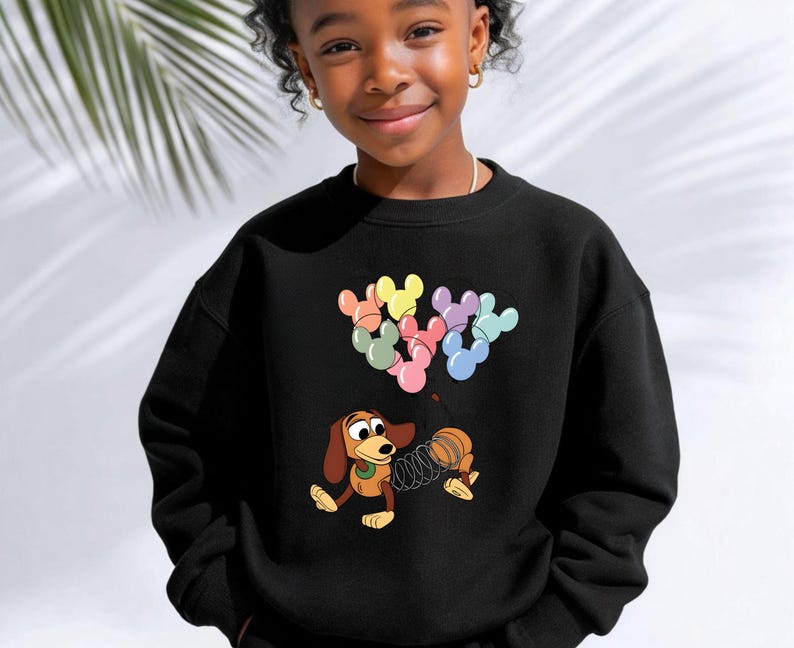 May include: Black sweatshirt featuring a cartoon image of Slinky Dog from Toy Story. The design includes colorful Mickey Mouse-shaped balloons. The sweatshirt is a crew neck style.