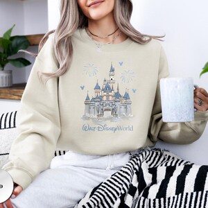 May include: A sand-coloured crewneck sweatshirt featuring a blue and white graphic illustration of a castle with the text Walt Disney World printed below. The design includes small firework and mouse ear icons surrounding the central castle image.