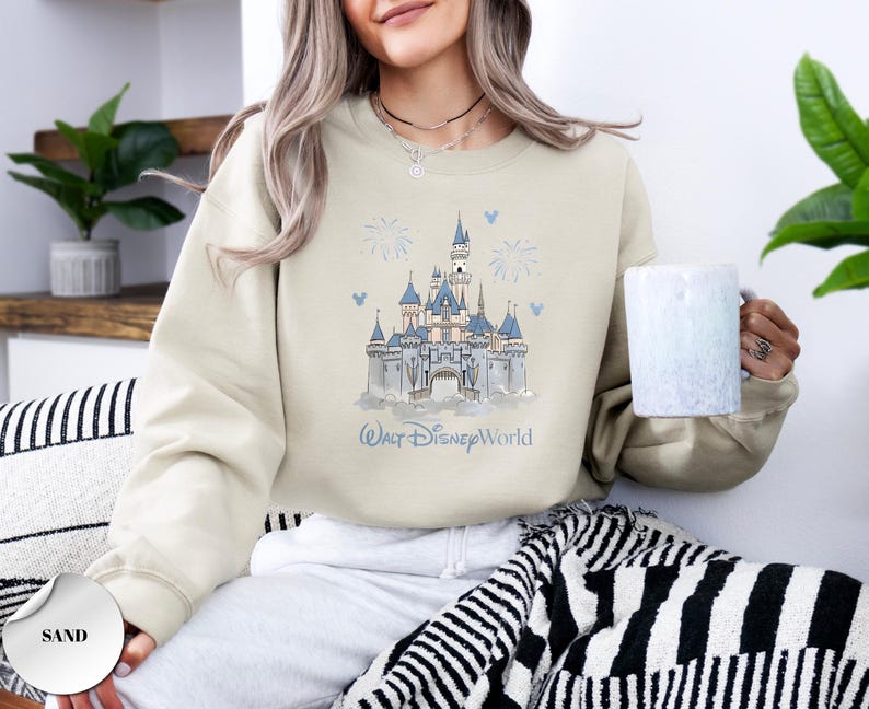 May include: A sand-coloured crewneck sweatshirt featuring a blue and white graphic illustration of a castle with the text Walt Disney World printed below. The design includes small firework and mouse ear icons surrounding the central castle image.