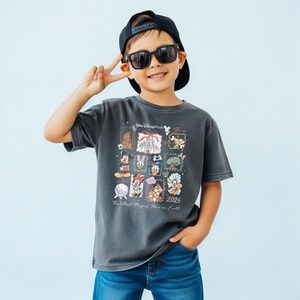 K&ouml;nnte beinhalten: Ein anthrazitfarbenes T-Shirt mit einem farbenfrohen Disney World-Design mit verschiedenen Charakteren und dem Text "2026 The Most Magical Place on Earth". Das T-Shirt wird von einer Person mit Sonnenbrille und einer schwarzen Baseballkappe getragen.