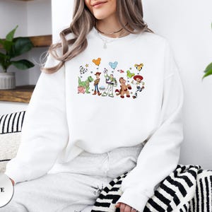 Op de afbeelding: Een wit sweatshirt met ronde hals voorzien van een kleurrijke grafische print van diverse geanimeerde speelgoedfiguren, waaronder een cowboy, een space ranger, een cowgirl, een dinosaurus, een varken en een paard met ballonnen.