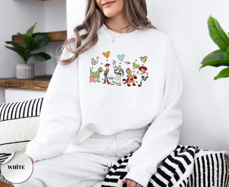 Op de afbeelding: Een wit sweatshirt met ronde hals voorzien van een kleurrijke grafische print van diverse geanimeerde speelgoedfiguren, waaronder een cowboy, een space ranger, een cowgirl, een dinosaurus, een varken en een paard met ballonnen.