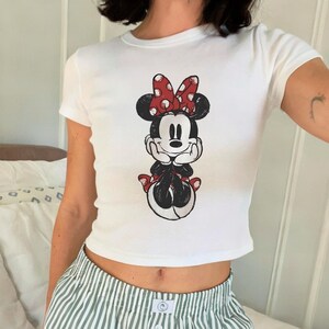 May include: White, short-sleeved crop top featuring a distressed graphic of Minnie Mouse. Minnie is depicted in black and white with a red polka-dot bow and accents. The shirt is a fitted style, suitable for casual wear.