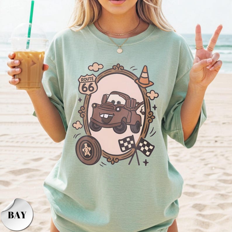 May include: A sage green t-shirt featuring a graphic print of a brown tow truck character inside an oval frame. The design includes a Route 66 sign, a traffic cone, a tire, and two checkered flags. The shirt has a crew neckline and short sleeves.