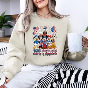 May include: A sand-colored crewneck sweatshirt featuring a graphic of Disney characters in patriotic attire, including Mickey, Minnie, Donald, Daisy, Goofy, and Pluto. The text reads 250 YEARS 1776 - 2026 with American flags and fireworks.
