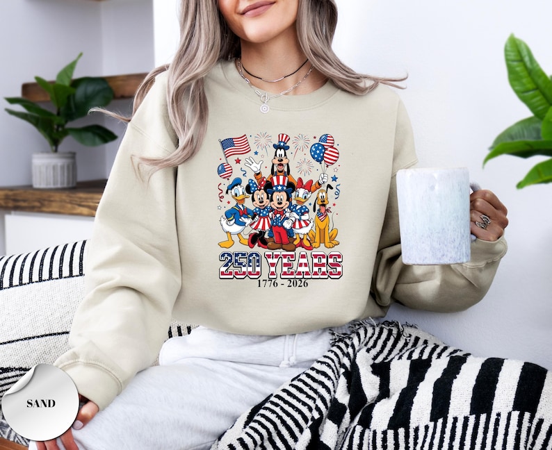 May include: A sand-colored crewneck sweatshirt featuring a graphic of Disney characters in patriotic attire, including Mickey, Minnie, Donald, Daisy, Goofy, and Pluto. The text reads 250 YEARS 1776 - 2026 with American flags and fireworks.