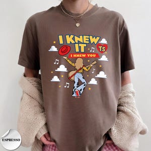 Puede incluir: Una camiseta marr&oacute;n con un estampado gr&aacute;fico con el texto I KNEW IT y I KNEW YOU. El dise&ntilde;o incluye una figura de dibujos animados con una guitarra, un sombrero rojo, un coraz&oacute;n rojo con TS, notas musicales, nubes blancas y estrellas amarillas.