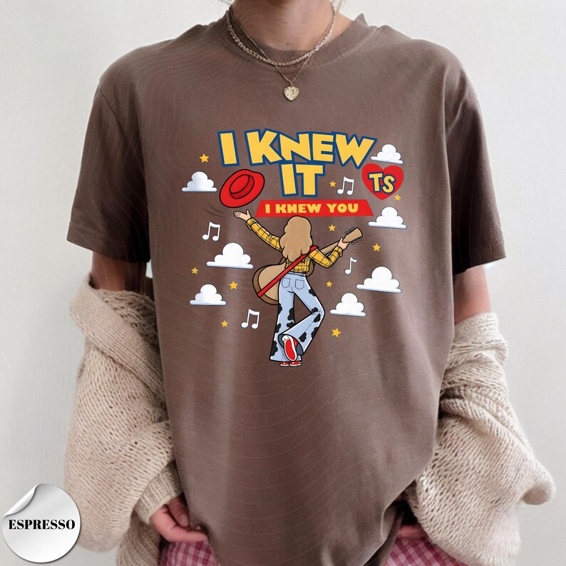 Puede incluir: Una camiseta marr&oacute;n con un estampado gr&aacute;fico con el texto I KNEW IT y I KNEW YOU. El dise&ntilde;o incluye una figura de dibujos animados con una guitarra, un sombrero rojo, un coraz&oacute;n rojo con TS, notas musicales, nubes blancas y estrellas amarillas.