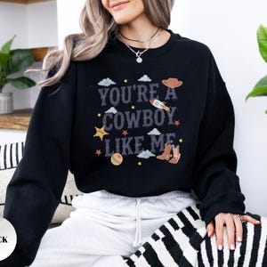 May include: A black crewneck sweatshirt featuring a graphic print with the text "YOU'RE A COWBOY LIKE ME" in a distressed font. The design includes illustrations of a rocket, a cowboy hat, a pair of boots, a ball, stars, and clouds.