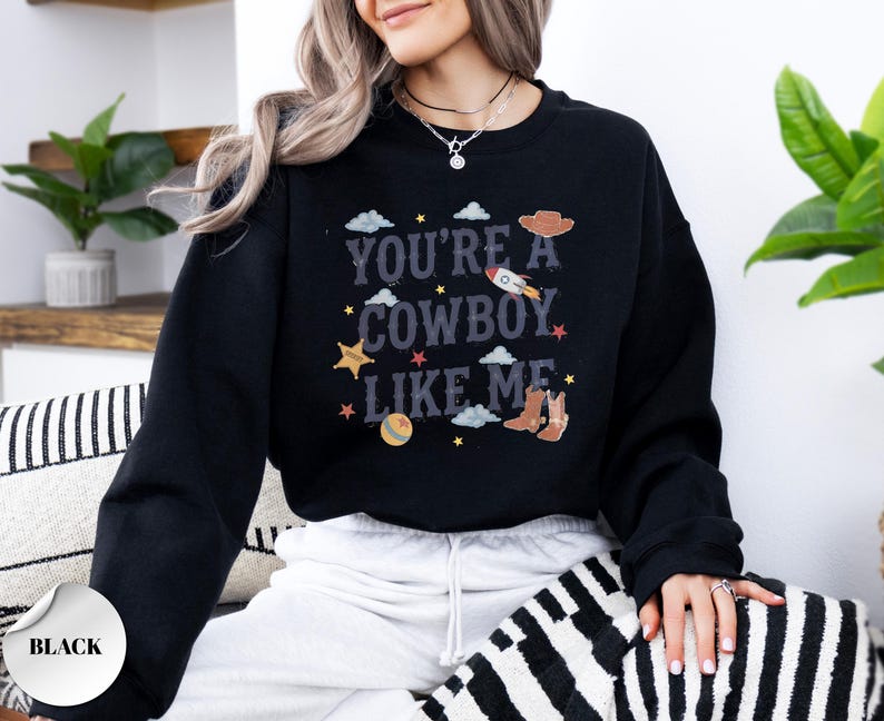 May include: A black crewneck sweatshirt featuring a graphic print with the text "YOU'RE A COWBOY LIKE ME" in a distressed font. The design includes illustrations of a rocket, a cowboy hat, a pair of boots, a ball, stars, and clouds.