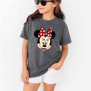 Op de afbeelding: Een grijs t-shirt met een Minnie Mouse-afbeelding. De afbeelding toont Minnie's gezicht met een rode strik met witte stippen. Het shirt heeft korte mouwen en wordt gecombineerd met een witte short. De persoon draagt een zwarte zonnebril.