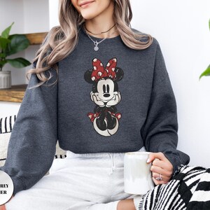 May include: Dark grey heather sweatshirt featuring a Minnie Mouse graphic. The graphic shows Minnie Mouse with a red bow with white polka dots. The sweatshirt is a crew neck style and has long sleeves. The text "DARK GREY HEATHER" is in a circle at the bottom left.