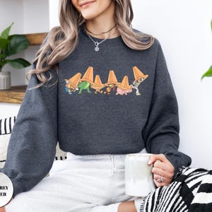 Op de afbeelding: Donkergrijze heather sweatshirt met een afbeelding van stripfiguren met oranje verkeerskegels. Het ontwerp bevat een dinosaurus, een varken en andere figuren. De sweatshirt heeft een ronde hals en lange mouwen.