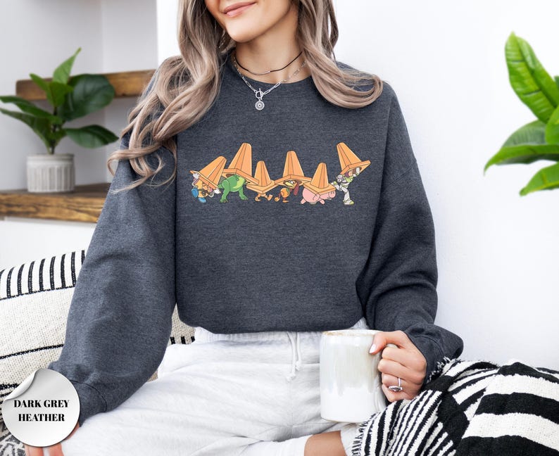 Op de afbeelding: Donkergrijze heather sweatshirt met een afbeelding van stripfiguren met oranje verkeerskegels. Het ontwerp bevat een dinosaurus, een varken en andere figuren. De sweatshirt heeft een ronde hals en lange mouwen.