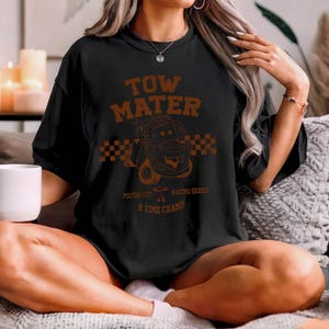 May include: A black short-sleeve t-shirt featuring a brown graphic print of the character Tow Mater. The design includes a checkered flag pattern, the text "TOW MATER", "PISTON CUP RACING SERIES", and "5 TIME CHAMP" in a distressed font style.