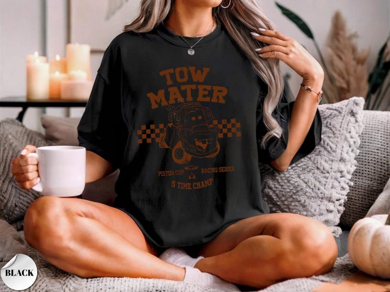 May include: A black short-sleeve t-shirt featuring a brown graphic print of the character Tow Mater. The design includes a checkered flag pattern, the text "TOW MATER", "PISTON CUP RACING SERIES", and "5 TIME CHAMP" in a distressed font style.