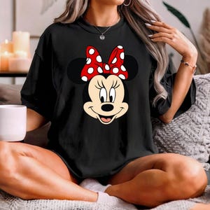 Op de afbeelding: Zwart T-shirt met een Minnie Mouse-ontwerp. Het ontwerp toont Minnie's gezicht met een rode strik met witte stippen. Het T-shirt heeft een ronde hals en is gemaakt van een zachte stof. Het is een casual kledingstuk.