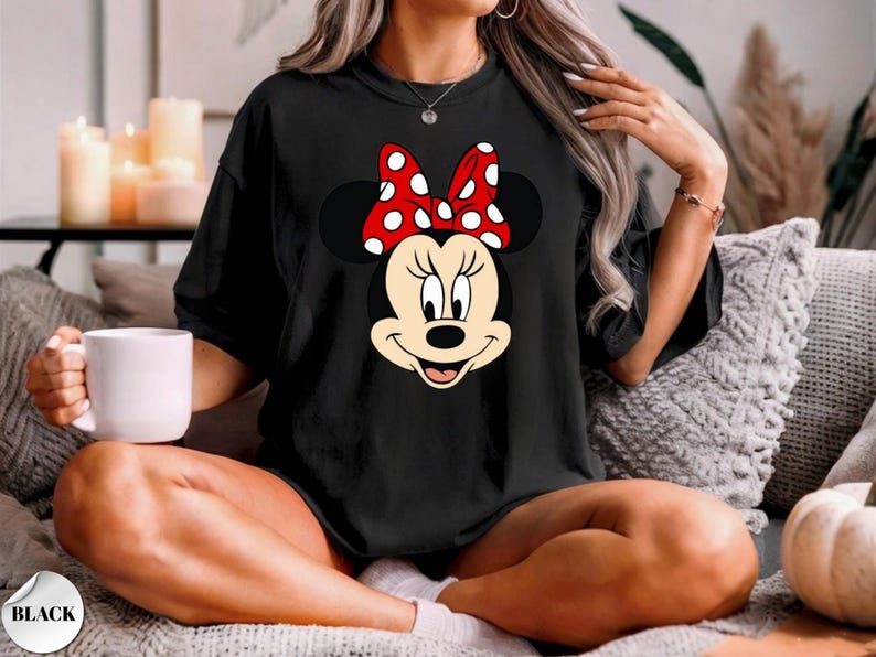 Op de afbeelding: Zwart T-shirt met een Minnie Mouse-ontwerp. Het ontwerp toont Minnie's gezicht met een rode strik met witte stippen. Het T-shirt heeft een ronde hals en is gemaakt van een zachte stof. Het is een casual kledingstuk.