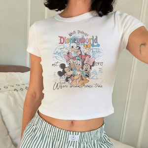 May include: White crop top featuring a colorful graphic of Disney characters and the text "Walt Disney's Disneyworld est. 1971 Where dreams come true." The shirt has short sleeves and a crew neck.