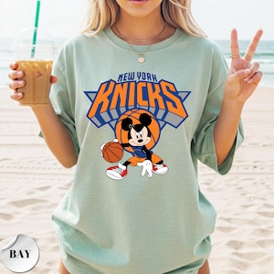 Puede incluir: Una camiseta de color verde salvia claro con un gr&aacute;fico de Mickey Mouse sosteniendo un bal&oacute;n de baloncesto. Sobre el personaje, el texto NEW YORK KNICKS aparece en letras naranjas y azules. El dise&ntilde;o incluye un bal&oacute;n de baloncesto.