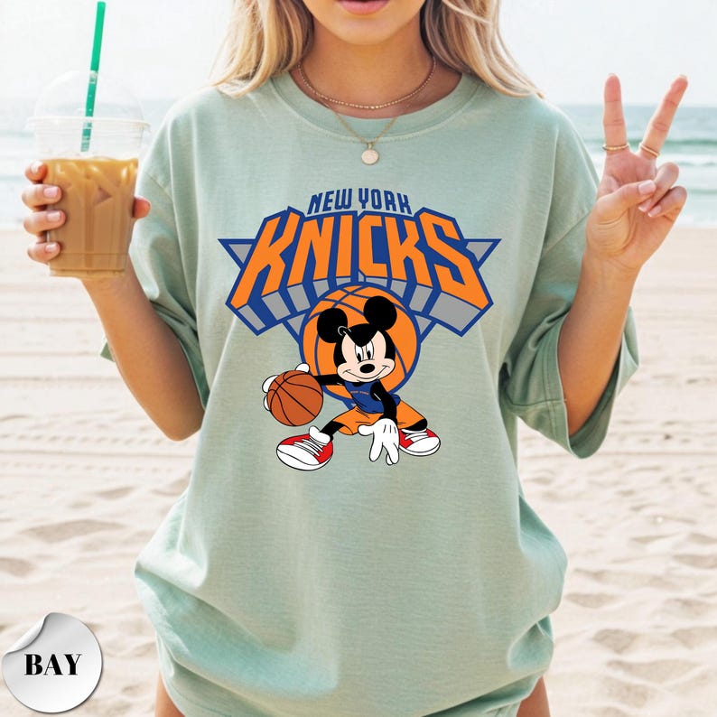 Puede incluir: Una camiseta de color verde salvia claro con un gr&aacute;fico de Mickey Mouse sosteniendo un bal&oacute;n de baloncesto. Sobre el personaje, el texto NEW YORK KNICKS aparece en letras naranjas y azules. El dise&ntilde;o incluye un bal&oacute;n de baloncesto.
