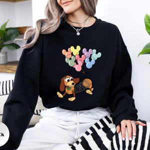 May include: Black sweatshirt featuring a cartoon illustration of Slinky Dog and Mickey Mouse-shaped balloons in pastel colors. The design includes a coiled spring and a playful, animated style. The sweatshirt is a solid black color.