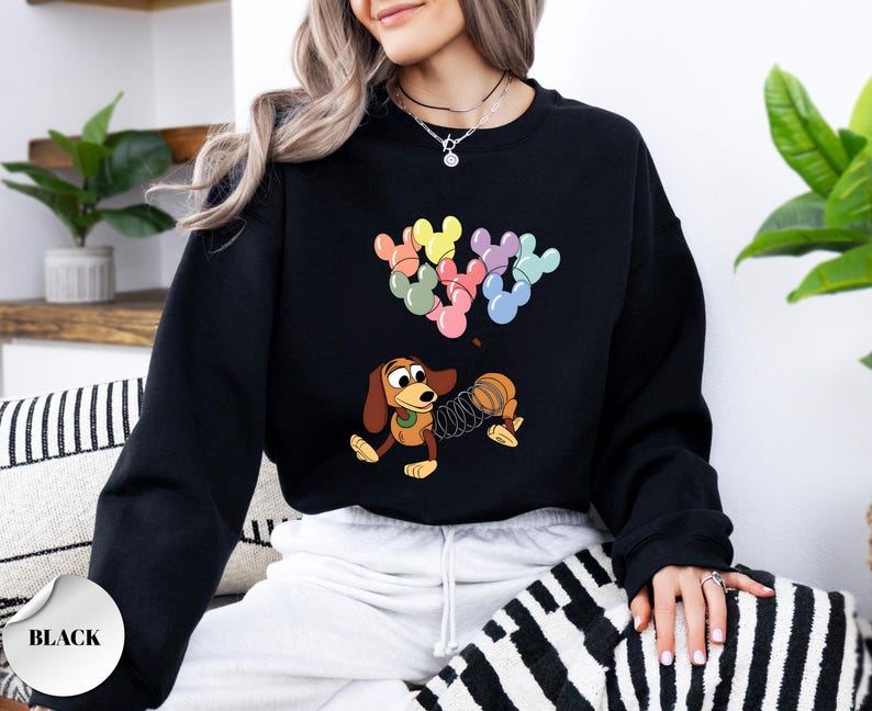 May include: Black sweatshirt featuring a cartoon illustration of Slinky Dog and Mickey Mouse-shaped balloons in pastel colors. The design includes a coiled spring and a playful, animated style. The sweatshirt is a solid black color.