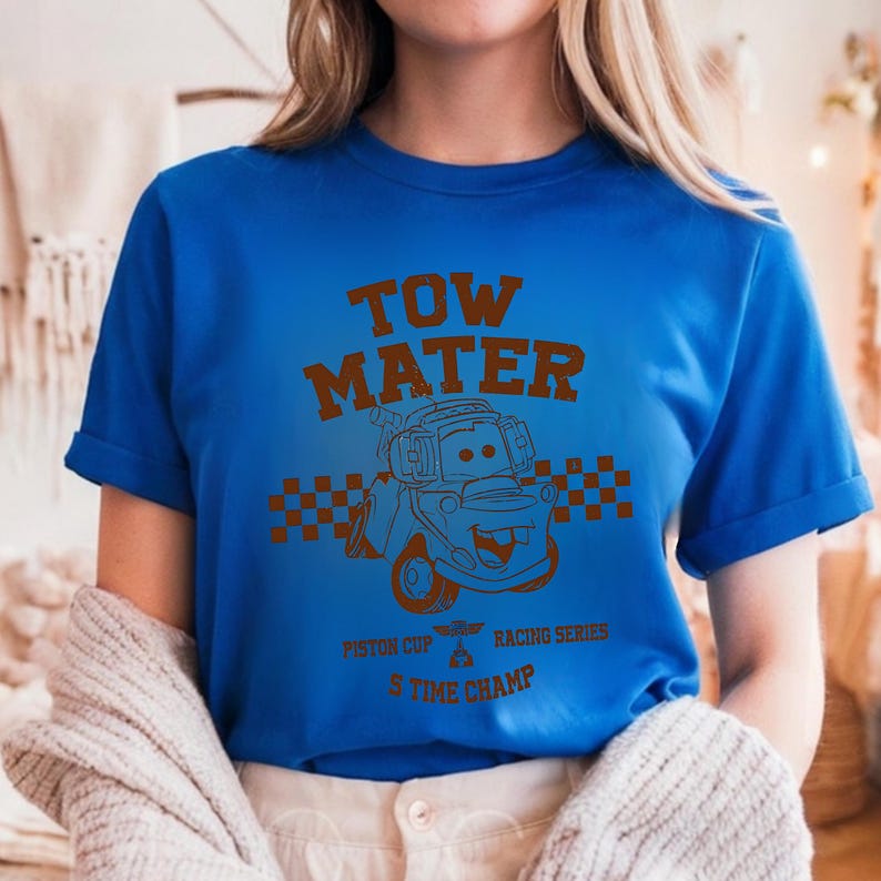 May include: A bright blue short-sleeved t-shirt featuring a brown graphic of the character Tow Mater. The design includes the text "TOW MATER", "PISTON CUP RACING SERIES", and "5 TIME CHAMP" with a checkered flag pattern behind the character.