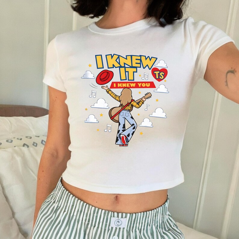 May include: A white short-sleeved t-shirt featuring a graphic print of a person with long hair holding a guitar, wearing a yellow shirt and cow-print trousers. The text reads I KNEW IT I KNEW YOU with a red hat, a red heart with TS, and clouds.