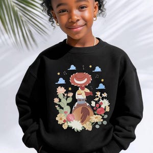 K&ouml;nnte beinhalten: Ein schwarzes Sweatshirt mit Rundhalsausschnitt und einem Grafikdruck auf der Brust, der eine Cowgirl mit braunem Hut auf einem braunen Pferd zeigt, umgeben von bunten Blumen, Kakteen, blauen Wolken und gelben Sternen.