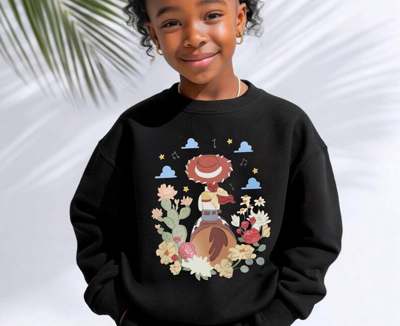 K&ouml;nnte beinhalten: Ein schwarzes Sweatshirt mit Rundhalsausschnitt und einem Grafikdruck auf der Brust, der eine Cowgirl mit braunem Hut auf einem braunen Pferd zeigt, umgeben von bunten Blumen, Kakteen, blauen Wolken und gelben Sternen.
