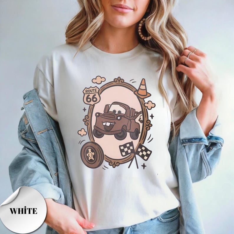 May include: A white t-shirt featuring a brown cartoon tow truck inside an oval frame. The graphic includes a Route 66 sign, a traffic cone, a tire, and two checkered flags. The design uses a muted color palette with brown, tan, and black accents.