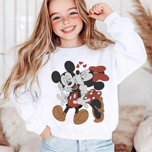 May include: White sweatshirt featuring a graphic of Mickey Mouse and Minnie Mouse kissing, with red hearts. Mickey is wearing red shorts and brown shoes, while Minnie has a red bow and polka dot skirt. The sweatshirt is a casual style.