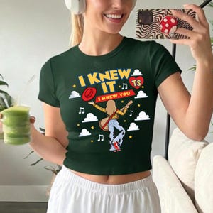May include: A dark green short-sleeved t-shirt featuring a graphic print with the text I KNEW IT I KNEW YOU and TS inside a red heart. The design includes a cartoon figure with a guitar, a red hat, musical notes, and white clouds on a dark background.