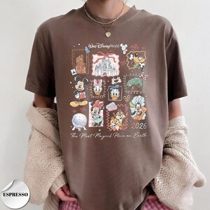 K&ouml;nnte beinhalten: Espressofarbenes T-Shirt mit einem farbenfrohen Disney World-Motiv. Das Design zeigt verschiedene Disney-Figuren, ein Schloss und den Text "2026 The Most Magical Place on Earth." Das Shirt hat kurze &Auml;rmel.