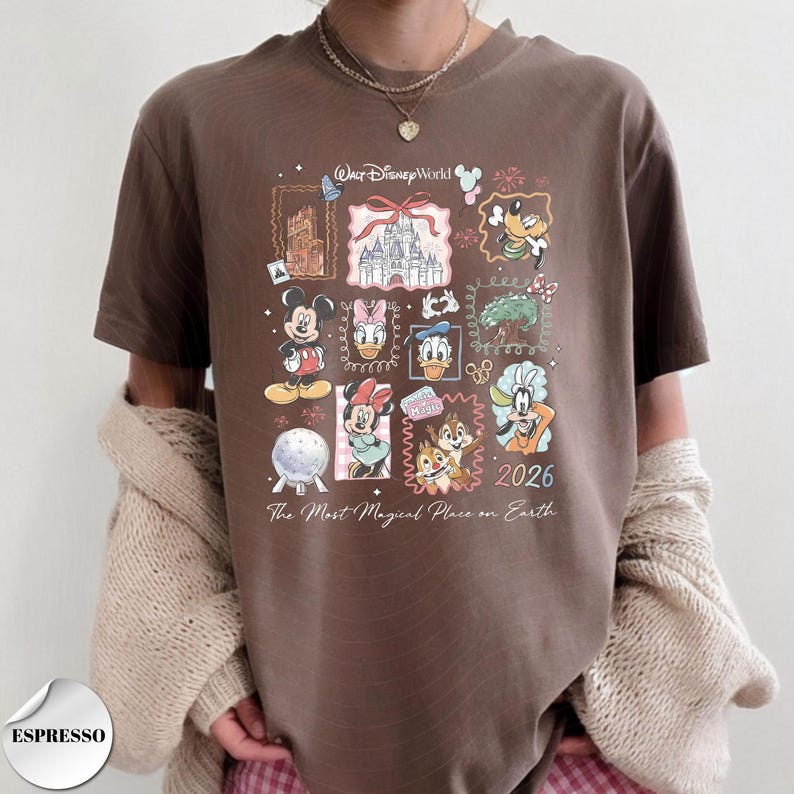 K&ouml;nnte beinhalten: Espressofarbenes T-Shirt mit einem farbenfrohen Disney World-Motiv. Das Design zeigt verschiedene Disney-Figuren, ein Schloss und den Text "2026 The Most Magical Place on Earth." Das Shirt hat kurze &Auml;rmel.
