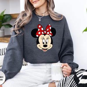 Puede incluir: Sudadera gris oscuro jaspeada con un gr&aacute;fico de Minnie Mouse. El gr&aacute;fico muestra la cara de Minnie con un lazo rojo con lunares blancos. El texto "DARK GREY HEATHER" est&aacute; impreso en la esquina inferior izquierda de la imagen.