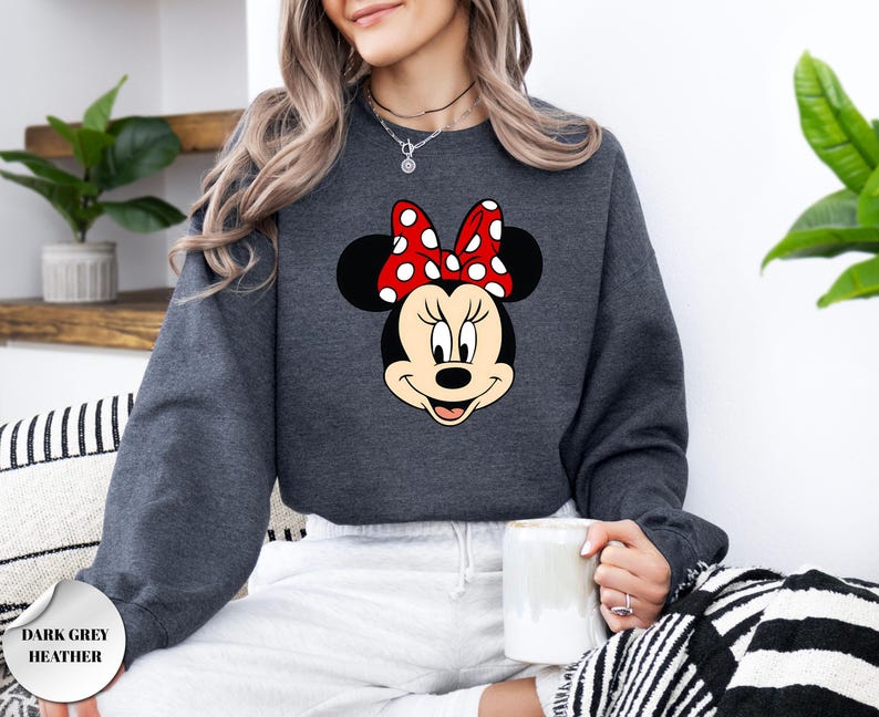 Puede incluir: Sudadera gris oscuro jaspeada con un gr&aacute;fico de Minnie Mouse. El gr&aacute;fico muestra la cara de Minnie con un lazo rojo con lunares blancos. El texto "DARK GREY HEATHER" est&aacute; impreso en la esquina inferior izquierda de la imagen.
