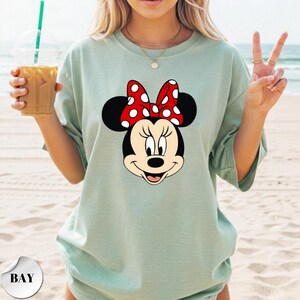 Op de afbeelding: Lichtgroen T-shirt met een Minnie Mouse-afbeelding. De afbeelding toont Minnie's gezicht met een rode strik met witte stippen. Het shirt heeft een relaxte pasvorm en korte mouwen. De achtergrond is een strandsc&egrave;ne.