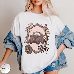 May include: A white t-shirt featuring a graphic print of a brown tow truck character inside an oval frame. The design includes a Route 66 sign, a traffic cone, a tire, and two checkered flags. The shirt is worn under a blue denim jacket.