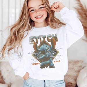 May include: A white long-sleeve sweatshirt featuring a graphic print of the blue character Stitch with arms crossed. The design includes the text STITCH in a distressed font, a diamond-shaped background, lightning bolts, and the text EXP 626.