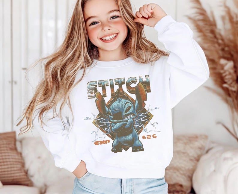 May include: A white long-sleeve sweatshirt featuring a graphic print of the blue character Stitch with arms crossed. The design includes the text STITCH in a distressed font, a diamond-shaped background, lightning bolts, and the text EXP 626.