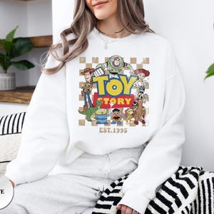 May include: A white crewneck sweatshirt featuring a graphic print of Toy Story characters including Woody, Buzz Lightyear, Jessie, Rex, Slinky Dog, Mr. Potato Head, and an alien. The design includes the text TOY STORY and EST. 1995 on a checkered background.