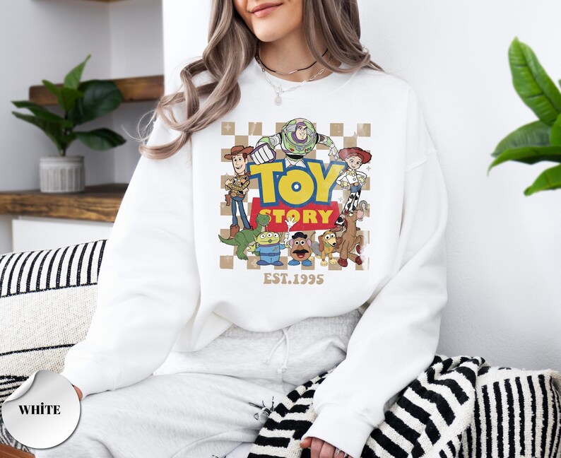 May include: A white crewneck sweatshirt featuring a graphic print of Toy Story characters including Woody, Buzz Lightyear, Jessie, Rex, Slinky Dog, Mr. Potato Head, and an alien. The design includes the text TOY STORY and EST. 1995 on a checkered background.