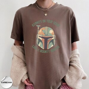 May include: A brown short-sleeve t-shirt featuring a graphic of a stylized helmet in shades of green, orange, and black. The text "KARMA'S ON YOUR SCENT" and "LIKE A BOUNTY HUNTER" is printed in green above and below the helmet illustration.