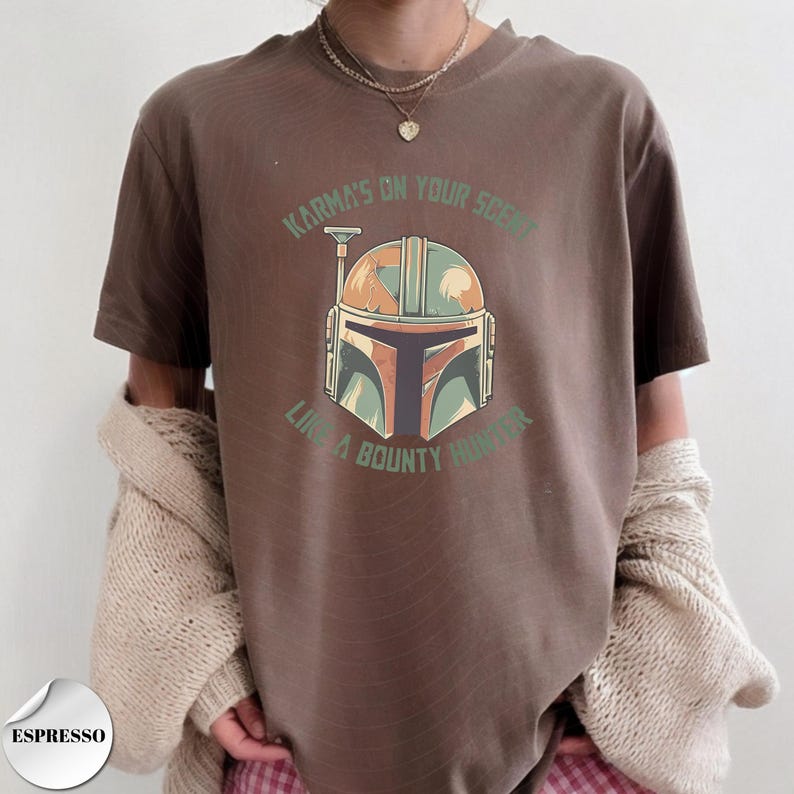 May include: A brown short-sleeve t-shirt featuring a graphic of a stylized helmet in shades of green, orange, and black. The text "KARMA'S ON YOUR SCENT" and "LIKE A BOUNTY HUNTER" is printed in green above and below the helmet illustration.