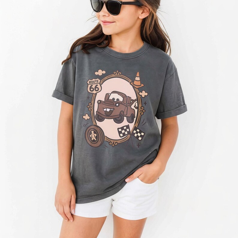 May include: A charcoal gray short-sleeve t-shirt featuring a graphic print of a brown tow truck character inside an oval frame. The design includes a Route 66 sign, a traffic cone, a tire, and checkered flags. The shirt is paired with white shorts.