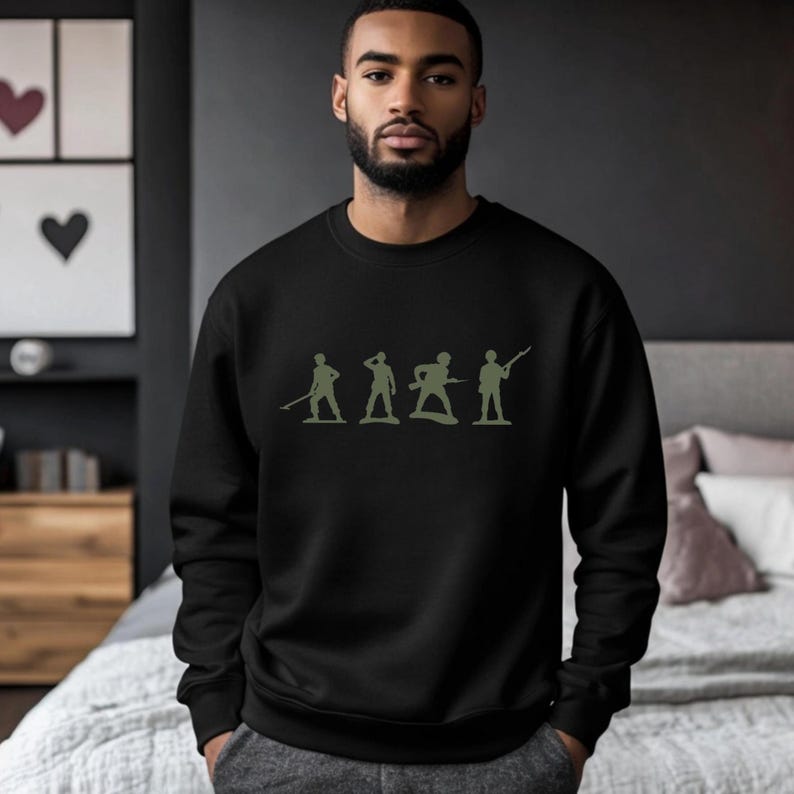 May include: Black sweatshirt featuring a green silhouette of toy soldiers. The design includes four figures in various poses, with one holding a rifle. The sweatshirt is made of a soft material and has a crew neck.