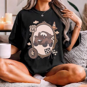 May include: A black t-shirt featuring a graphic print of a brown tow truck character inside an oval frame. The design includes a Route 66 sign, a traffic cone, a tire, and two checkered flags. The text Route 66 is printed on the sign.