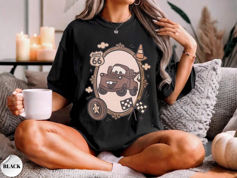 May include: A black t-shirt featuring a graphic print of a brown tow truck character inside an oval frame. The design includes a Route 66 sign, a traffic cone, a tire, and two checkered flags. The text Route 66 is printed on the sign.